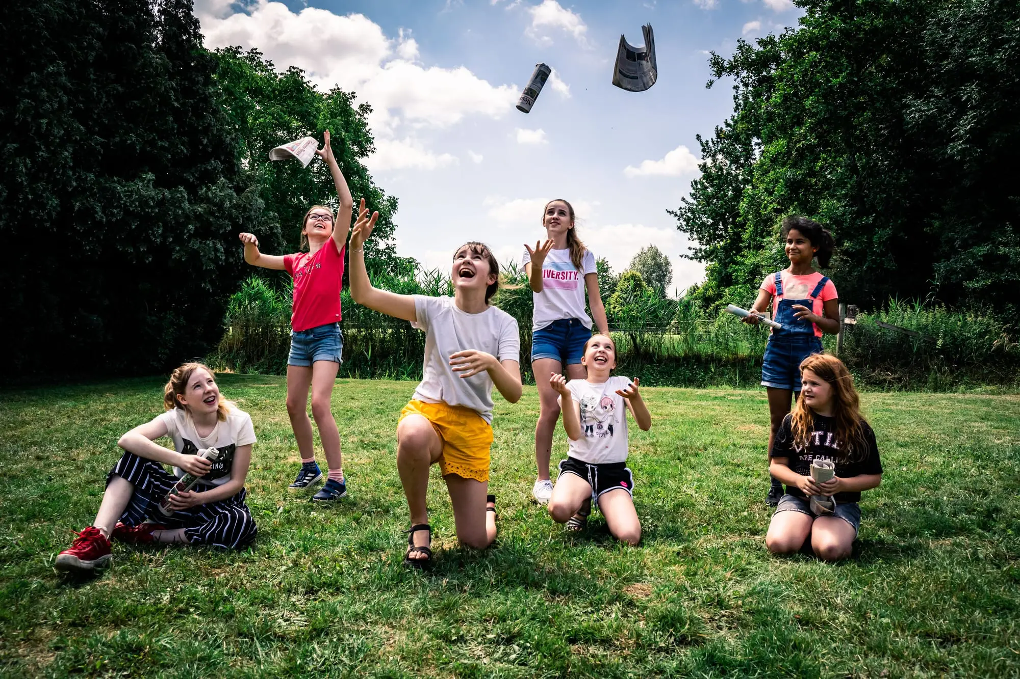 Een groep leerlingen speelt buiten in het gras, kranten vliegen door de lucht